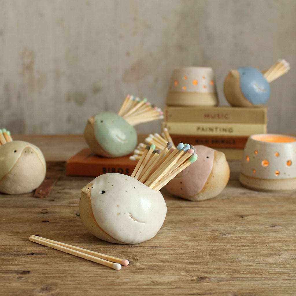Close up of white bird match pot with pastel matches in front of other pastel birds, books and tealights
