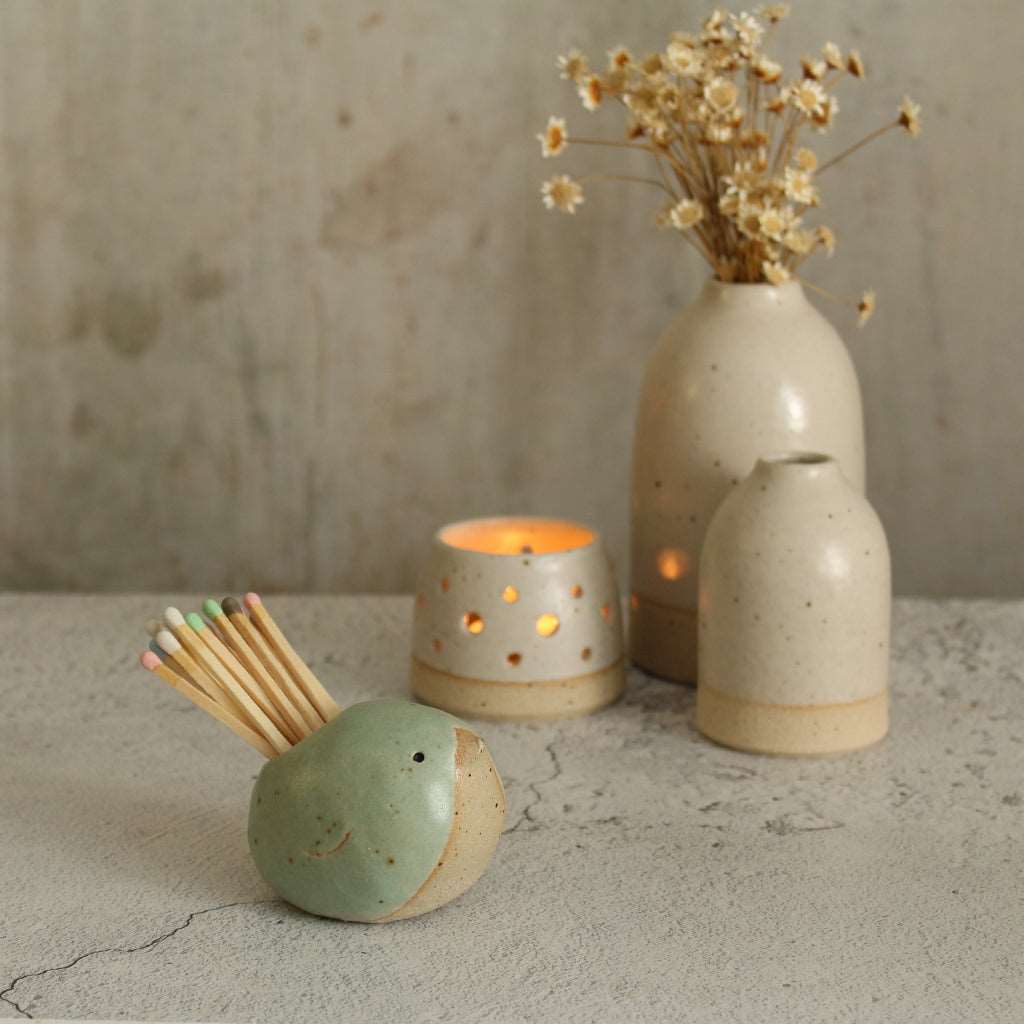 Green bird with pastel matches in front of a lit white tealight and white vases of flowers