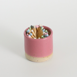 Pink ceramic match striker pot with multicoloured pastel wooden matches on a white background