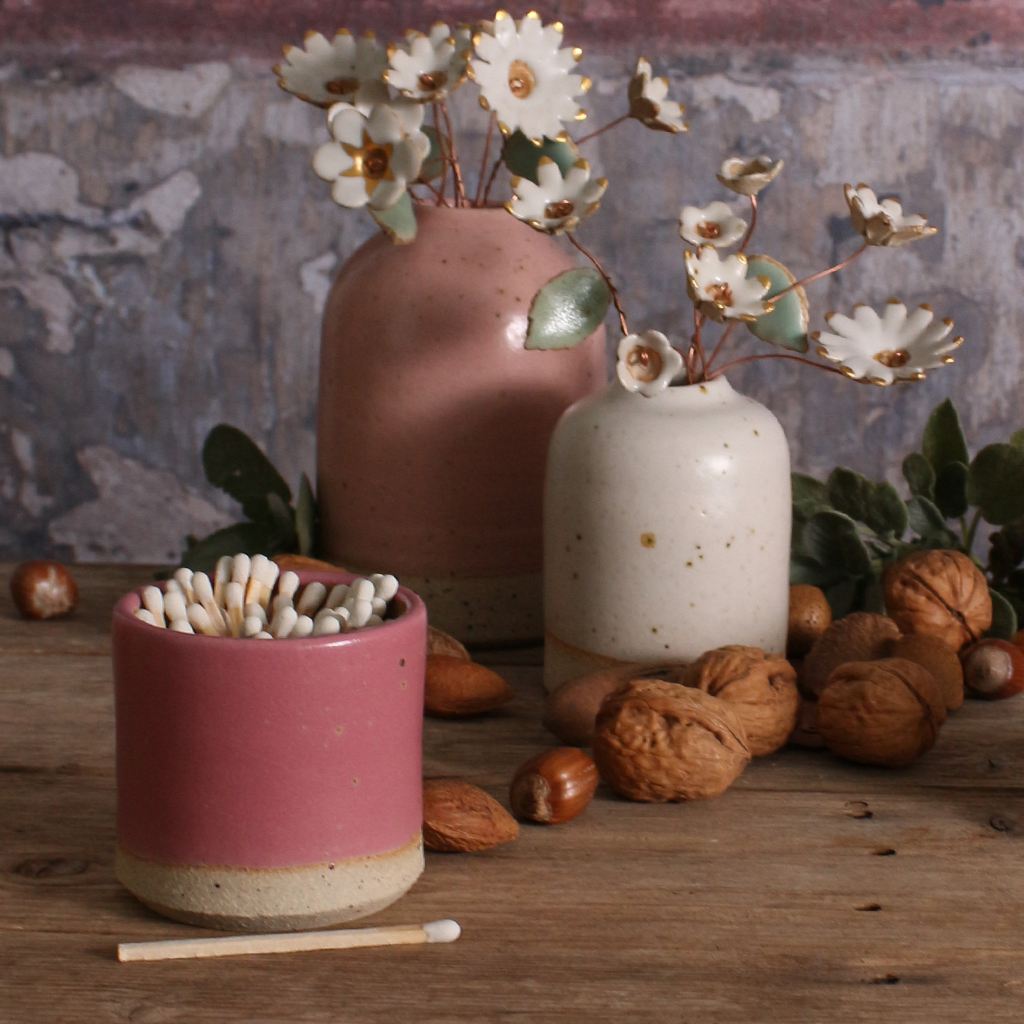 A fuchsia match striker pot with two ceramic pots with plants on a wooden surface.