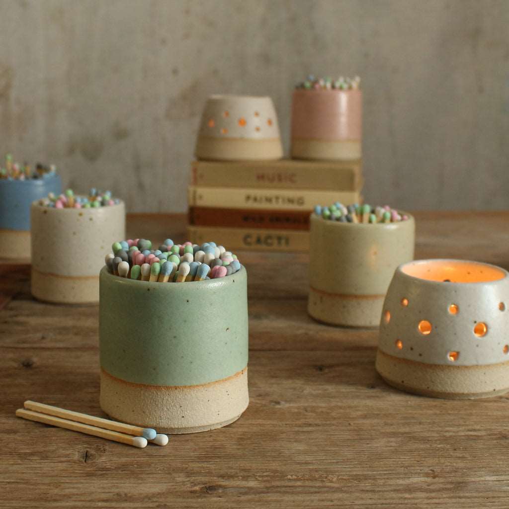 Close up of green match pot with pastel matches next to lit tealight in front of oter pastel match pots