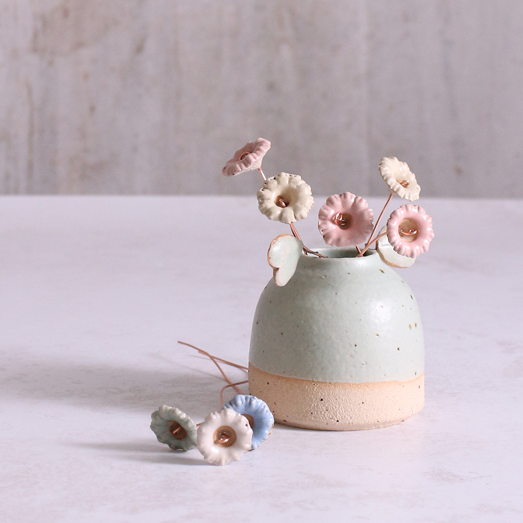 A bouquet of ceramic wildflower stems in pastel shades of pink, blue, and white, artfully arranged in a rustic ceramic vase and some laid next to the vase.