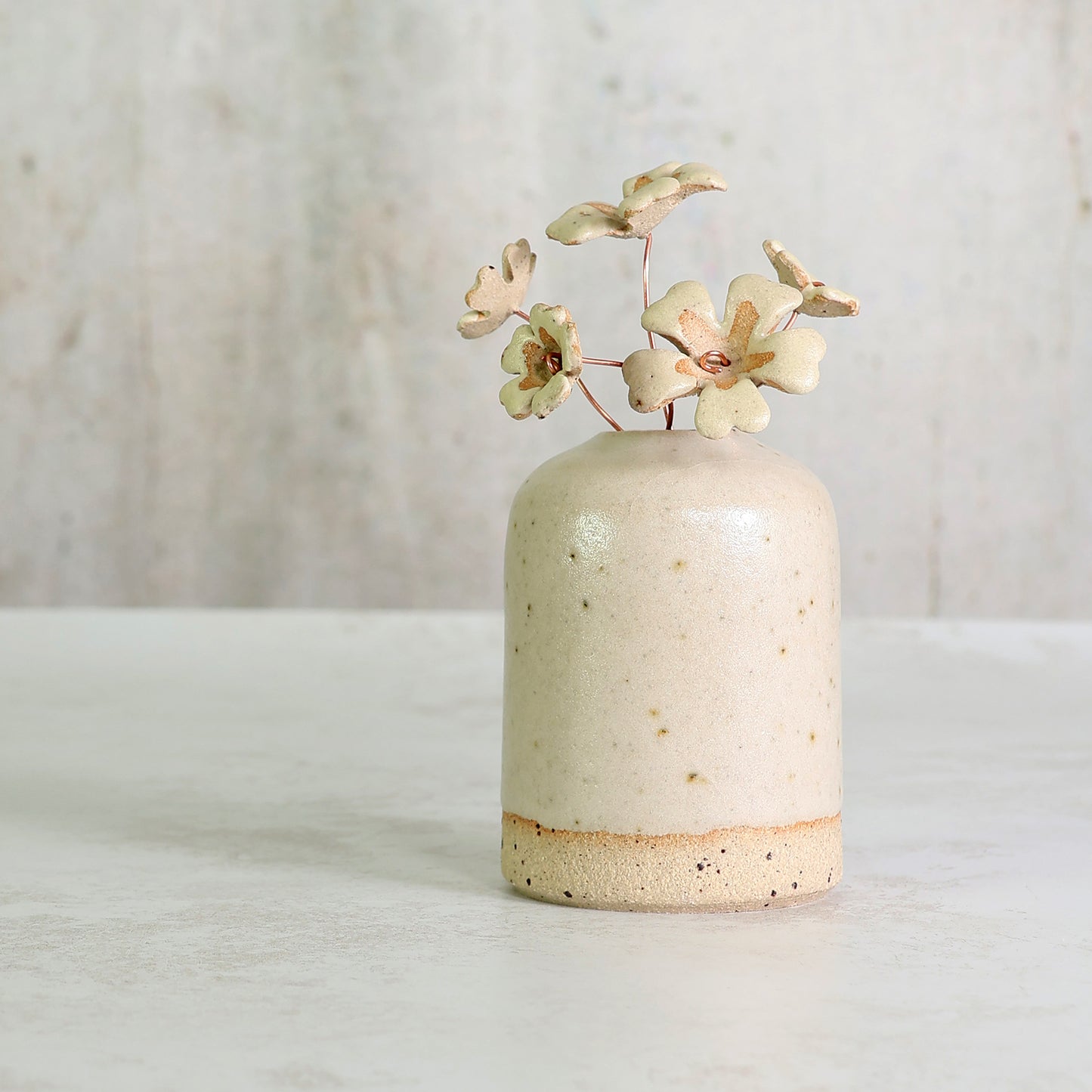 Small ceramic vase with speckled texture holding ceramic primrose flowers on a light background