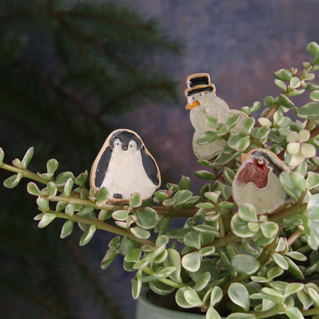Three small ceramic figurines of a penguin, a snowman, and a bird on a plant with a blurred background.
