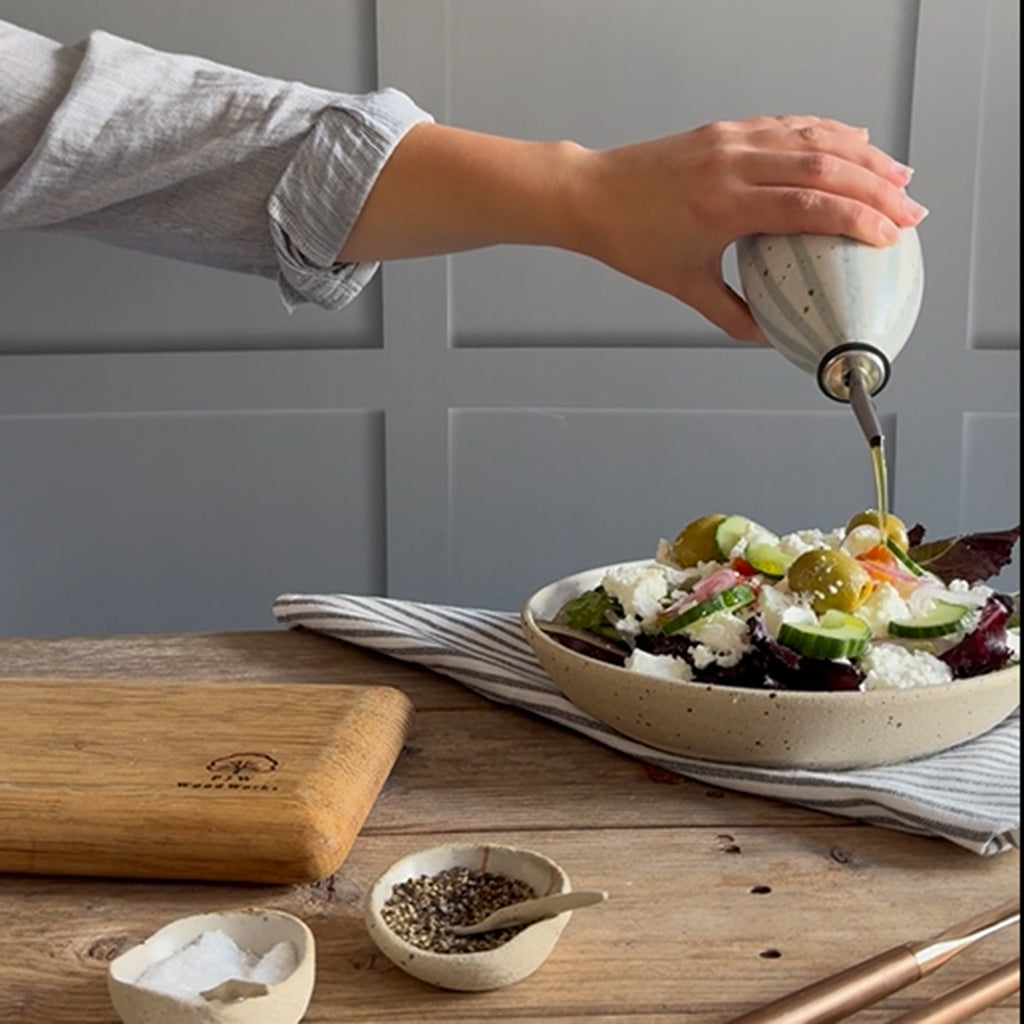 A person's hand pours olive oil from a handmade ceramic oil pourer with a blue and white stripe design into a fresh salad bowl on a wooden table, with a wooden cutting board and small handmade bowls nearby.