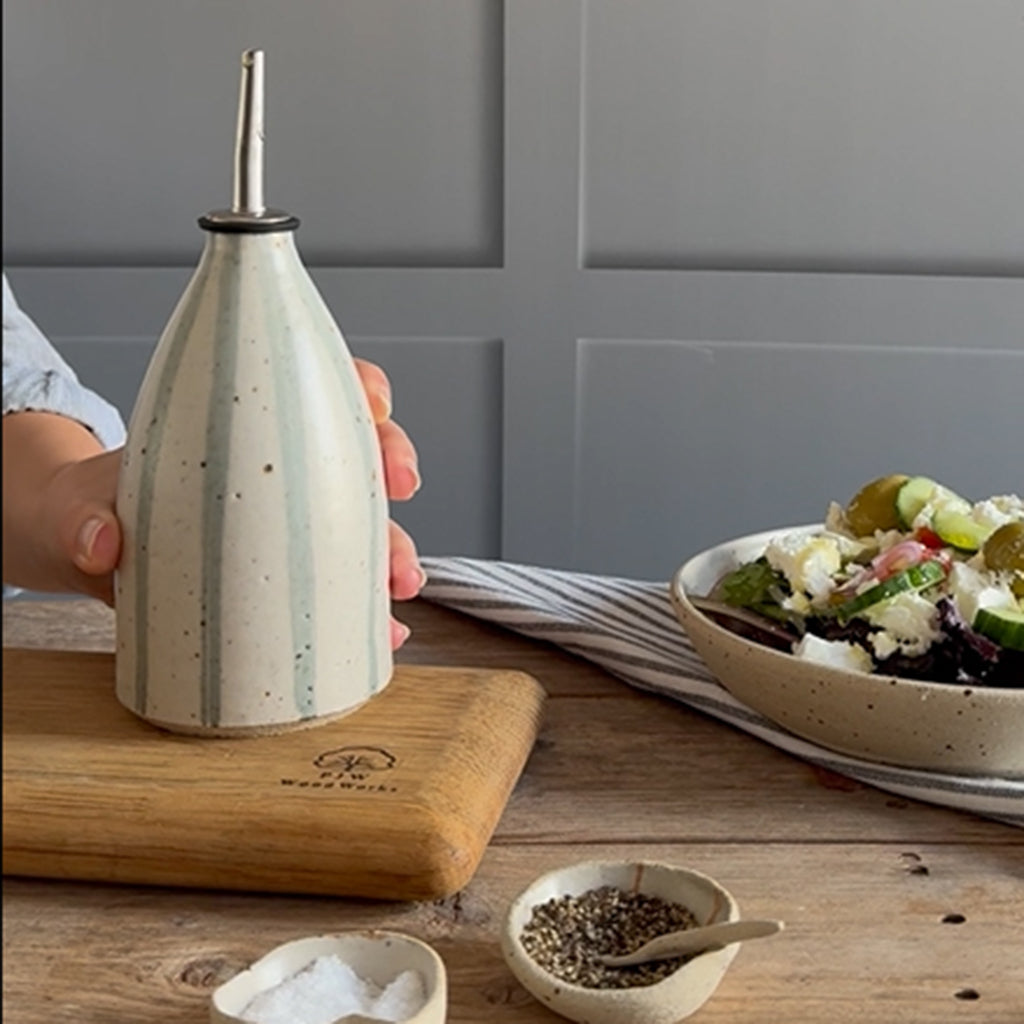 A ceramic oil dispenser with a blue and white stripe design and a stainless steel pour spout, held by a hand on a wooden cutting board next to a salad bowl.