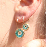 A women's ear wearing gold hoop earrings with round teal-coloured charms made from ceramic and glass. 