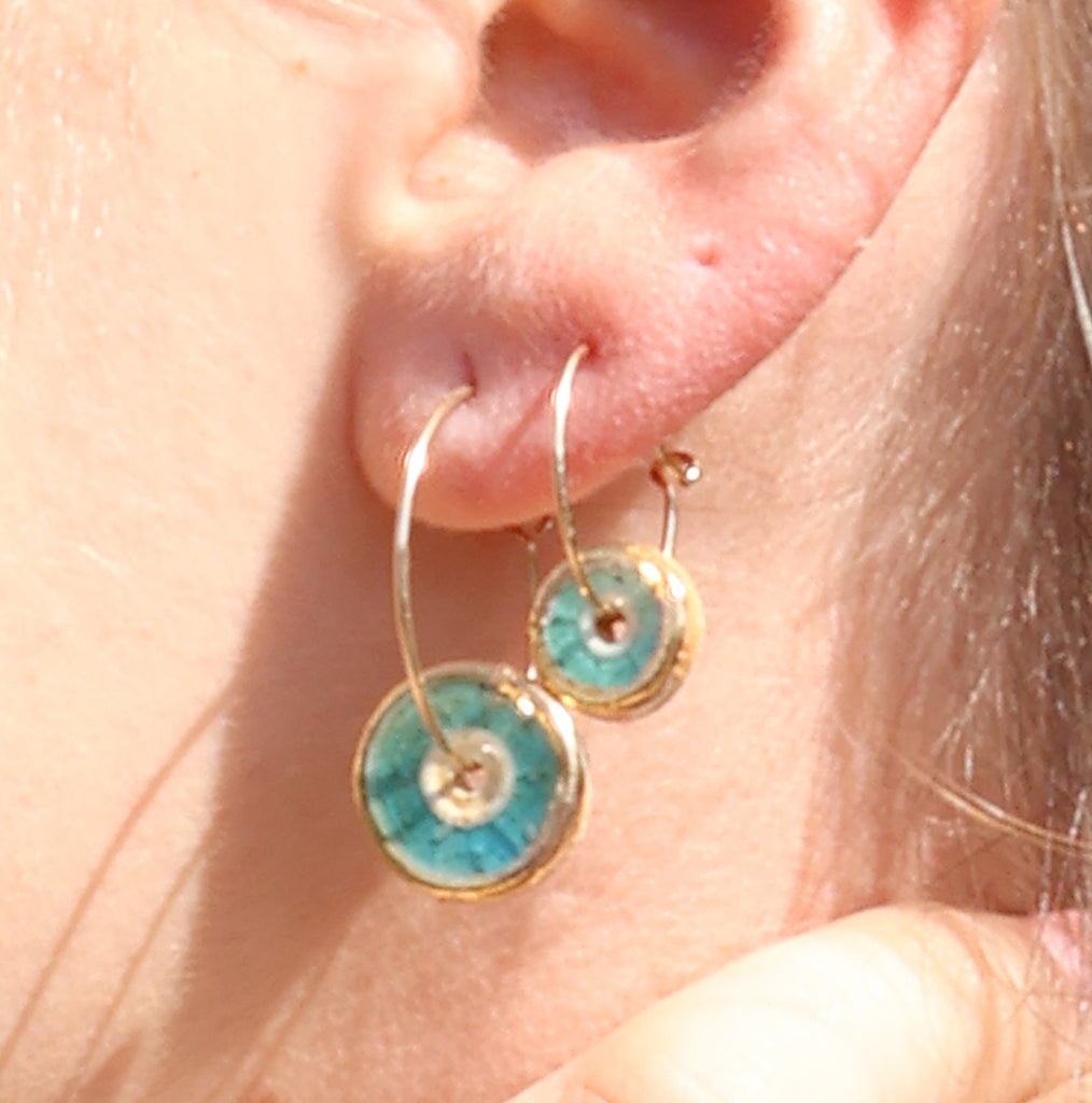 A women's ear wearing gold hoop earrings with round teal-coloured charms made from ceramic and glass. 