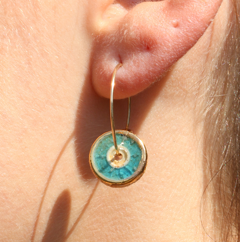 A women's ear wearing a gold fill hoop earring with a round teal-coloured charm made from ceramic and glass. 