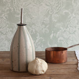 A handmade ceramic oil pourer with a stainless steel pour spout stands on a rustic wooden table next to a copper saucepan and a whole garlic bulb. 