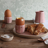 A handmade ceramic pink stripy egg cup holding a soft boiled egg sits on a pink plate of toast. A matching egg cup and small jug sit in the background. 