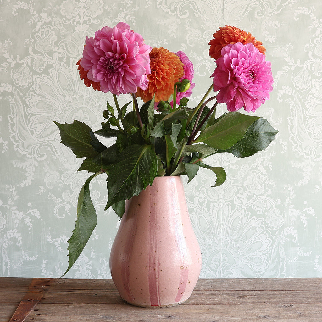 A pink striped ceramic vase filled with pink and orange dahlia flowers and green leaves, set on a wooden table against a pale damask wallpaper background.