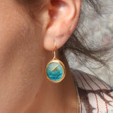 A close-up of a women's ear wearing a ceramic and glass teal and white oval drop earring with gold detailing. 