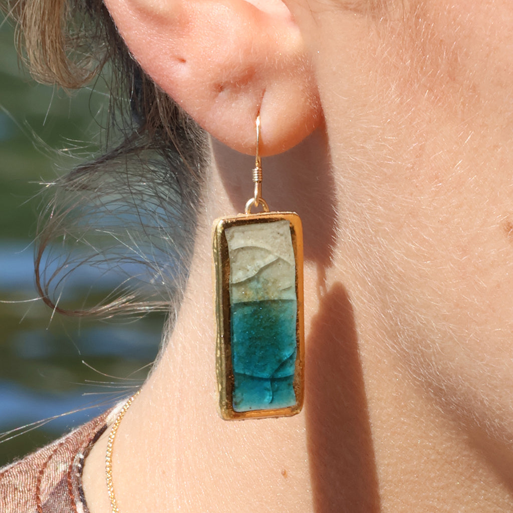 A close-up of a women's ear wearing a ceramic rectangular drop earring made from ceramic and teal and white glass with gold detailing. 