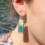 A close-up of a women's ear wearing a ceramic rectangular drop earring made from ceramic and teal and white glass with gold detailing. 