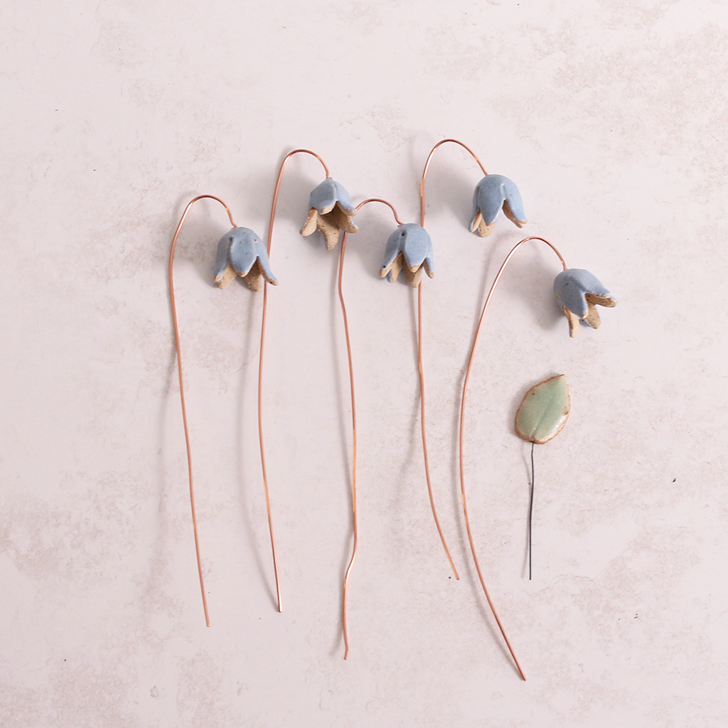 Handcrafted ceramic bluebell flower stems with soft glazes lay on a table. Each flower features a flexible copper wire stem for easy adjustment.