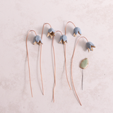 Handcrafted ceramic bluebell flower stems with soft glazes lay on a table. Each flower features a flexible copper wire stem for easy adjustment.