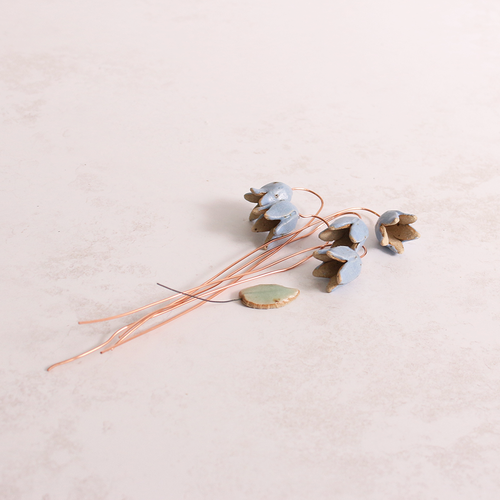 Handcrafted ceramic bluebell flower stems with soft glazes lay on a table. Each flower features a flexible copper wire stem for easy adjustment.