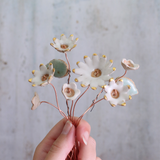 Hand holding a decorative arrangement of ceramic flowers with gold accents against a light gray background.