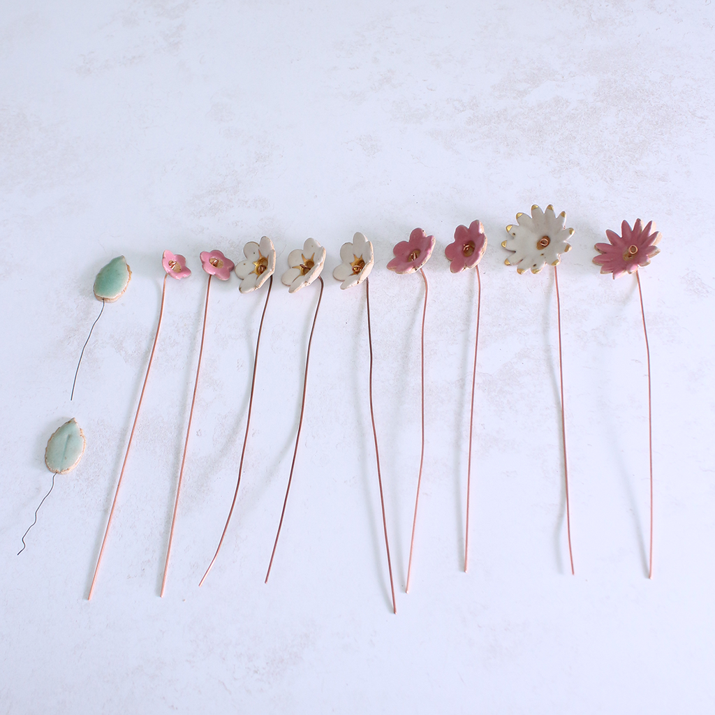 Handmade ceramic flower bouquet in white, pink, and fuchsia with gold-painted petals and copper wire stems displayed spread out on a table.