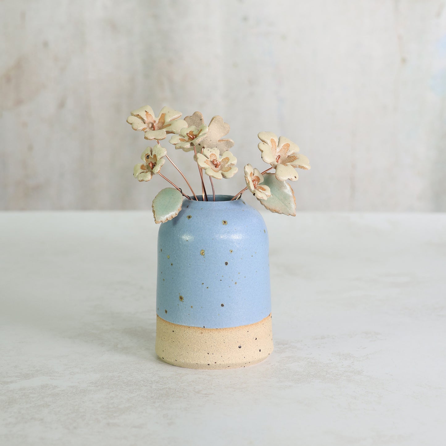 Blue ceramic vase with exposed clay bottom holding handmade ceramic flowers on copper wire on  a light background