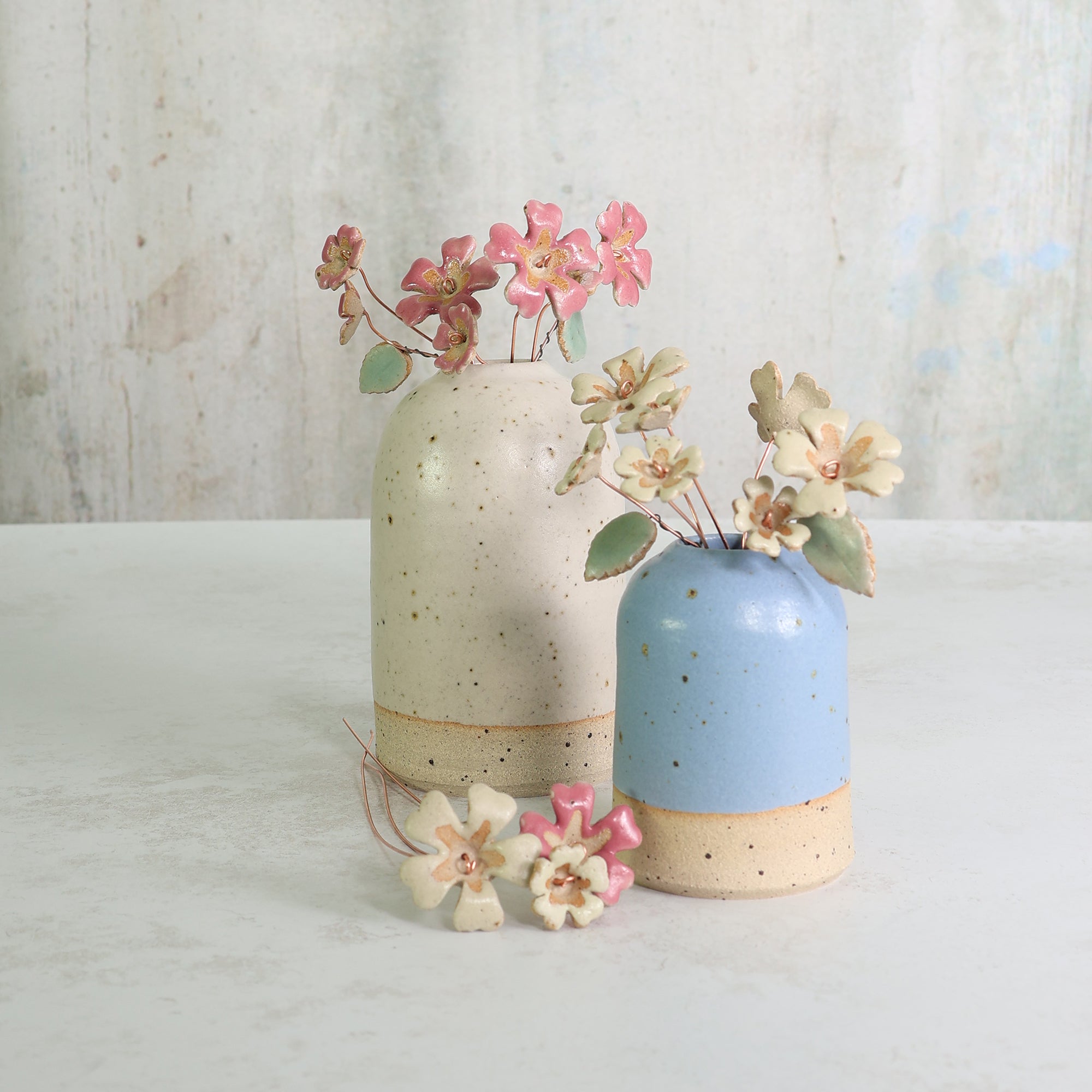 Two ceramic vases, one white and one blue, with handmade primrose flowers in pink and yellow on a light background