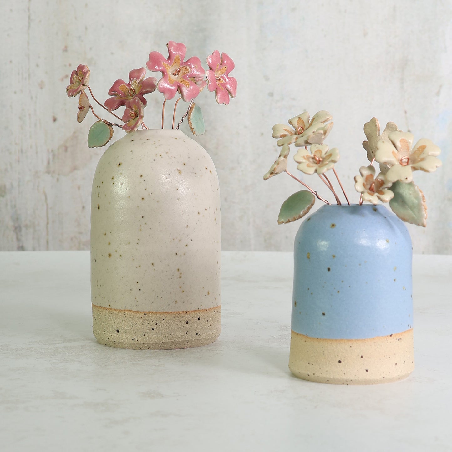 Two ceramic vases with speckled textures, one white and one blue, each containing small ceramic floral arrangements.