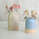 Two ceramic vases with speckled textures, one white and one blue, each containing small ceramic floral arrangements.