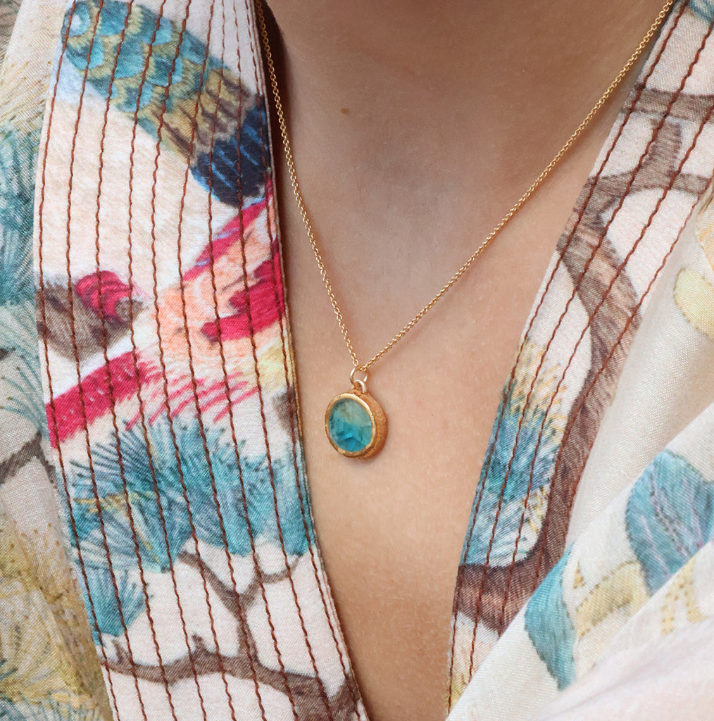 A women wearing a delicate gold chain necklace with an small round teal ceramic and glass pendant with gold detailing. 