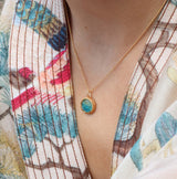 A women wearing a delicate gold chain necklace with an small round teal ceramic and glass pendant with gold detailing. 