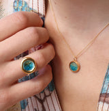 A close-up of a women wearing a delicate gold chain necklace with an small round teal ceramic and glass pendant with gold detailing, with a matching ring. 