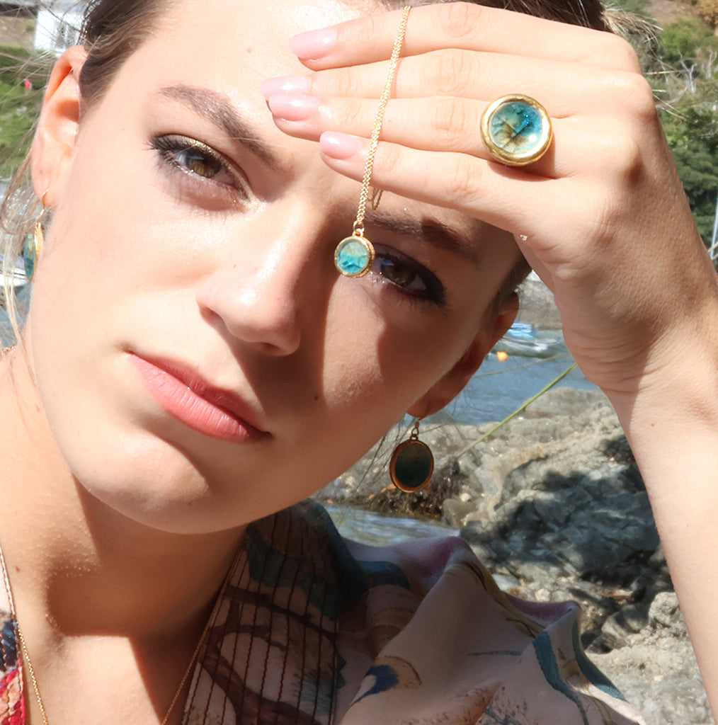A women wears a teal ceramic and glass ring with gold detailing, with matching oval drop earrings and a small round pendnant necklace. 