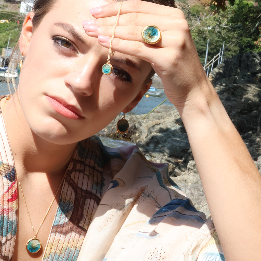 A woman outdoors wearing gold jewellery, including a round teal-blue pendant necklace made from ceramic and glass with matching drop earrings, ring and necklace with a coastal rocky background.