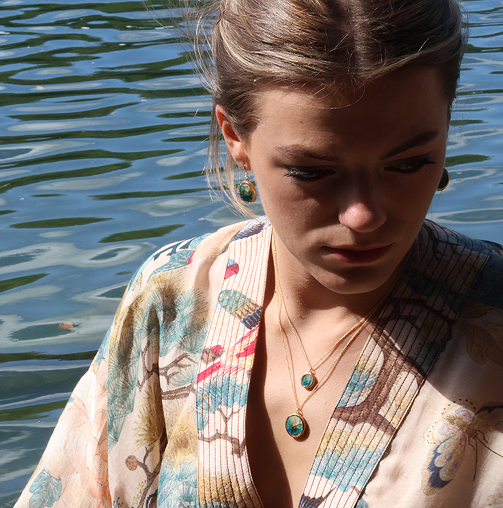 A women wears a ceramic and teal glass round pendant necklace with gold detailing, with a matching large round pendant and oval drop earrings, set against an ocean background. 