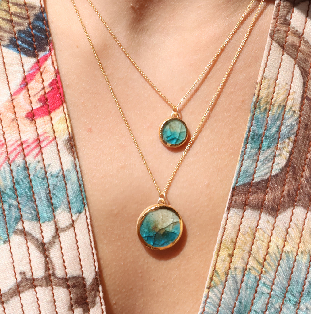 A close-up lifestyle shot of women wearing two delicate gold chain necklaces, one with a small round pendant and one with a larger round pendant made from ceramic and glass with gold detailing around the rim. The glass is blue, white and brown. 
