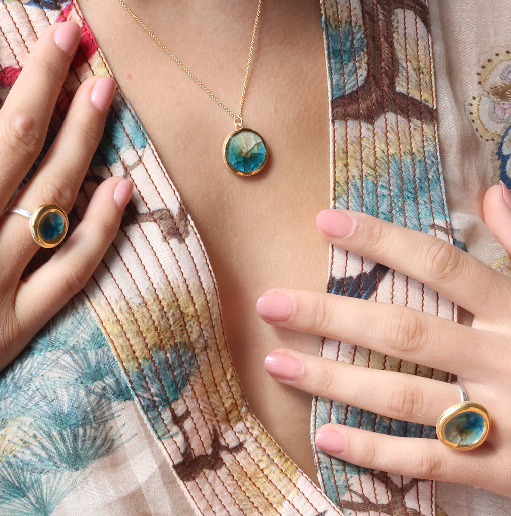 A women wearing a gold pendant necklace with a round charm made from ceramic and teal glass with gold detailing, with two matching rings. 