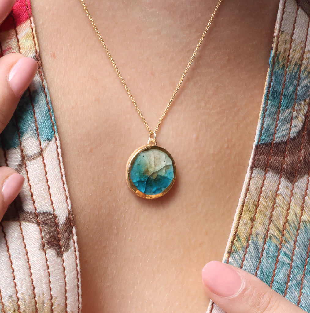 A close-up lifestyle shot of women wearing a gold chain necklace with an round pendant made from ceramic and glass with gold detailing around the rim. The glass is blue, white and brown. 