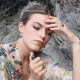 A women wearing matching teal ceramic and glass jewellery with gold detailing, set against a rocky shoreline background. She wears a ring, a round pendant necklace and oval drop earrings. 