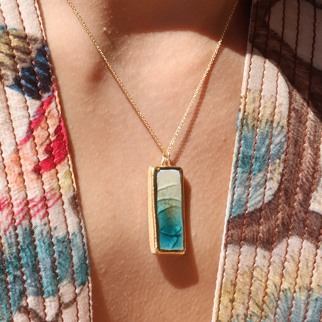 A women wears a rectangle drop pendant necklace made from ceramic and teal coloured glass with gold detailing. 