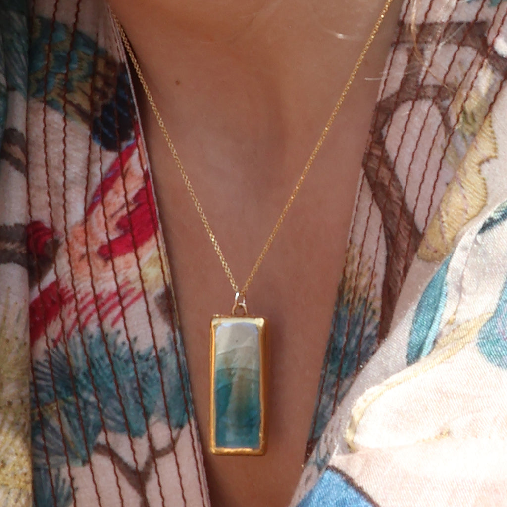 A women wears a rectangle drop pendant necklace made from ceramic and teal coloured glass with gold detailing. 
