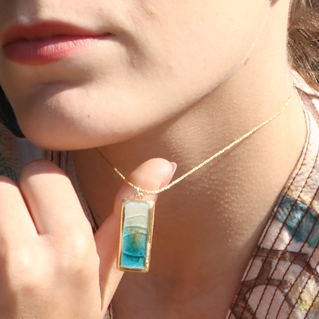 A women wears a rectangle drop pendant necklace made from ceramic and teal coloured glass with gold detailing. 
