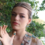 A woman wears a gold necklace and matching gold earrings with teardrop pendants made from ceramic and teal coloured glass with gold detailing. 