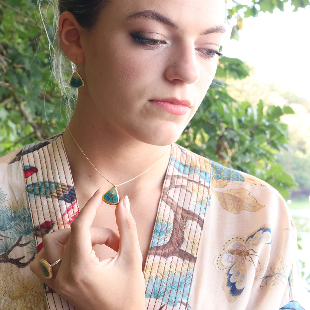 A women wears a delicate gold chain necklace with a teardrop-shaped pendant made from ceramic and teal coloured glass, with matching earrings and ring. 