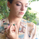 A women wears a delicate gold chain necklace with a teardrop-shaped pendant made from ceramic and teal coloured glass, with matching earrings and ring. 