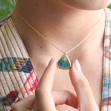 A women wears a delicate gold chain necklace with a teardrop-shaped pendant. The pendant is made from ceramic and glass in teal, white and brown with gold detailing, held gently between two fingers. 