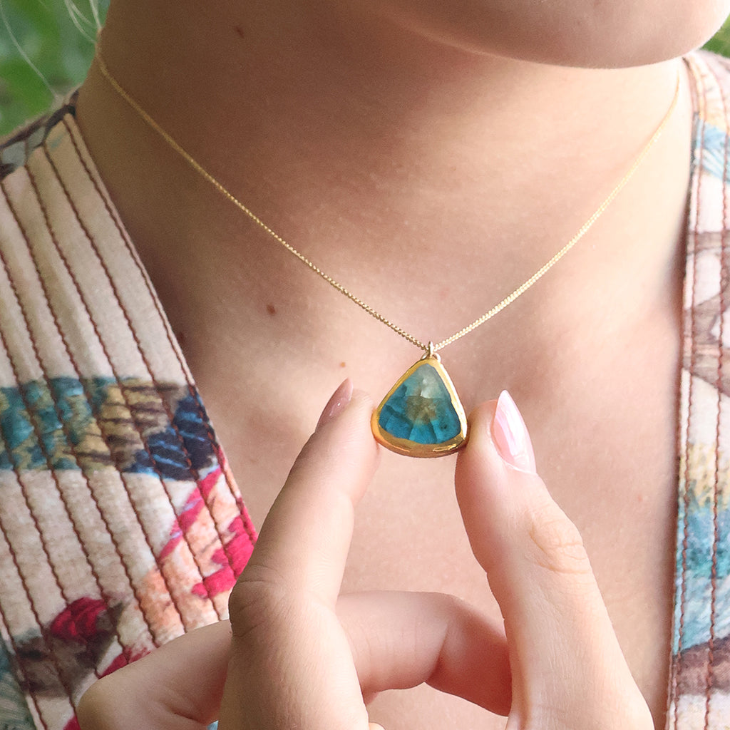 A women wears a delicate gold chain necklace with a teardrop-shaped pendant. The pendant is made from ceramic and glass in teal, white and brown with gold detailing, held gently between two fingers. 