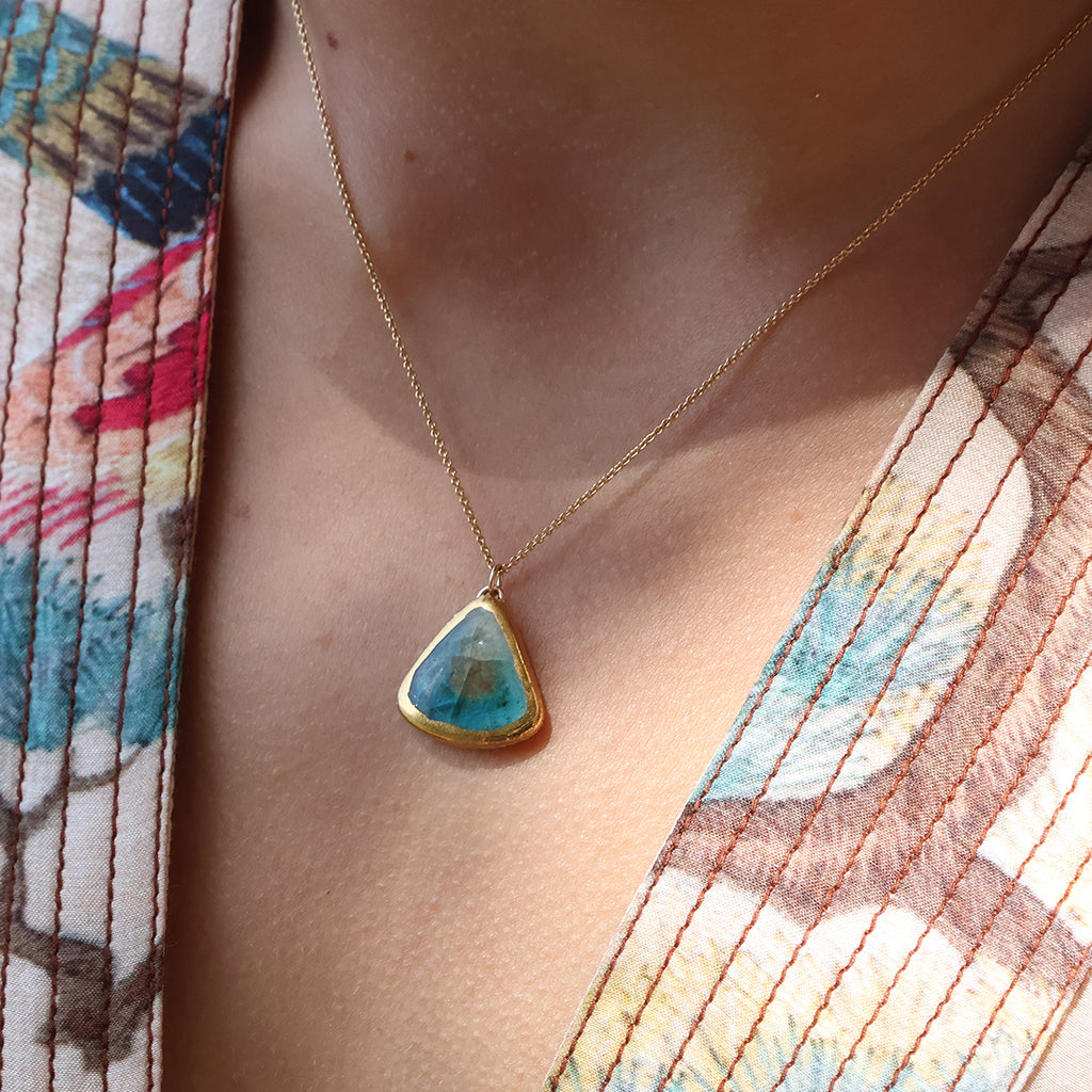 A women wears a gold necklace with a teardrop pendant made from a ceramic and teal coloured glass. 