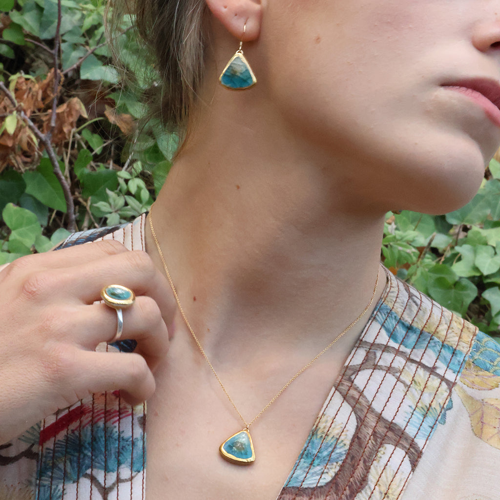 A women wears a delicate gold chain necklace with a teardrop-shaped pendant made from ceramic and teal coloured glass, with matching earrings and ring. 