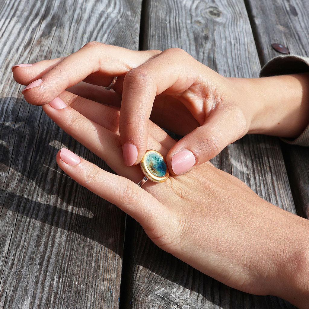 A close-up of a person's hands resting on a wooden surface, wearing a sterling silver ring with ceramic, glass and gold charms. The glass is teal, white and brown. 
