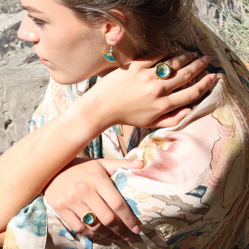 A women wearing ceramic, glass and gold rings and a matching teardrop earring, each made with teal, white and brown glass. 